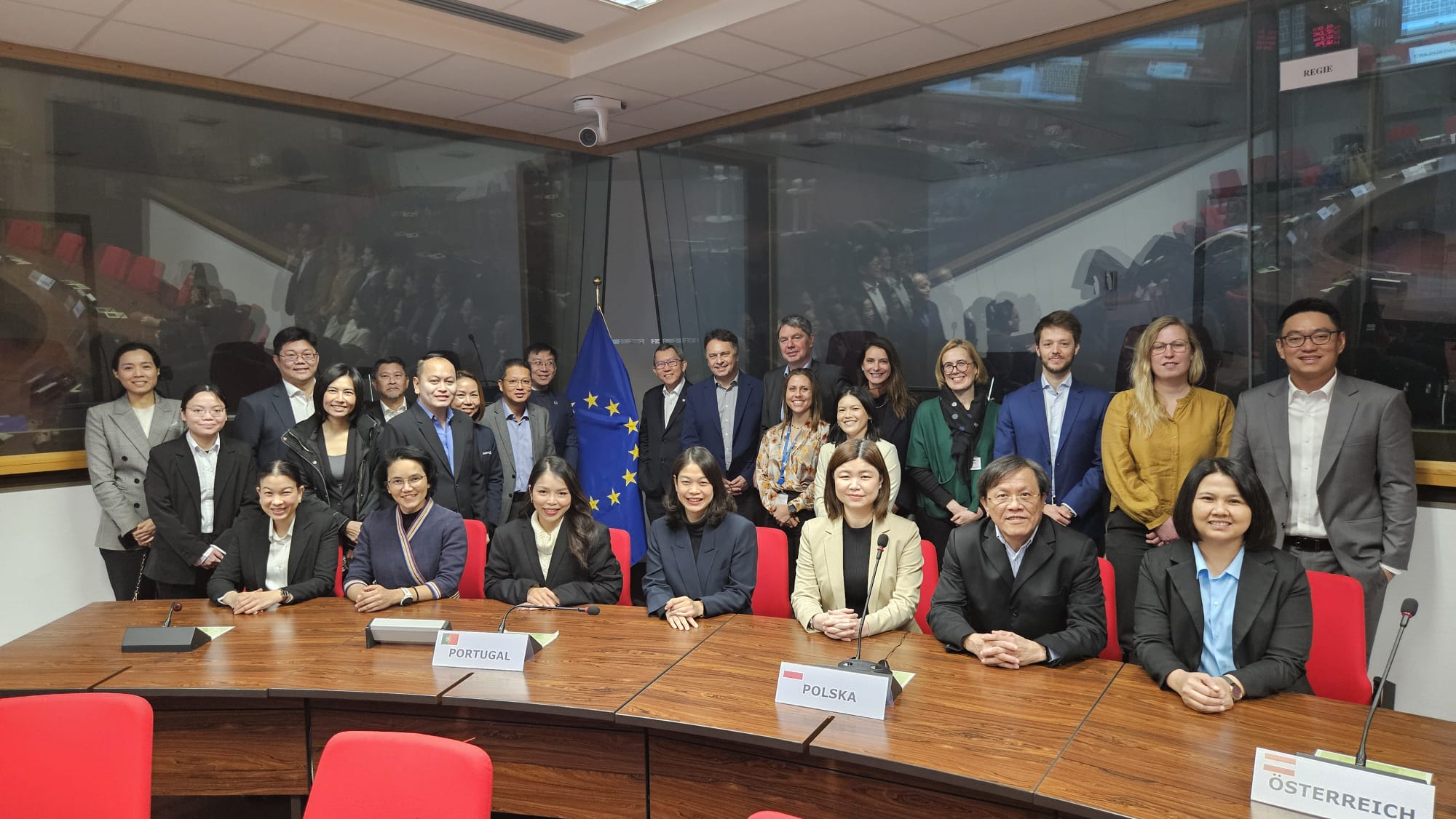 Ein Gruppenfoto von ca. 25 Personen in einem Konferenzraum. Sieben Personen sitzen an einem Konferenztisch mit Mikrofonen, die restlichen Personen stehen dahinter. Im Hintergrund ist die EU-Flagge zu sehen. 