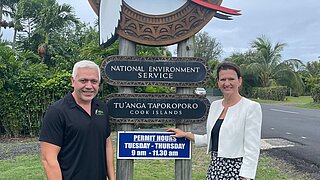 Zwei Personen stehen vor einem grossen Holzschild mit einem Vogel-Logo und der Aufschrift "National Environment Service" sowie "Tu’anga Taporoporo – Cook Islands".