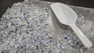 Nahaufnahme von zerkleinertem transparentem Plastik mit blauen Partikeln, verteilt auf einer Metalloberfläche. Rechts unten liegt eine weiße Kunststoffschaufel.