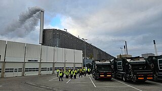 Der Parkplatz eines Fabrikgeländes mit einigen Personen in gelben Warnwesten und drei großen schwarzen Fahrzeugen. Im Hintergrund ein hoher qualmender Schornstein.