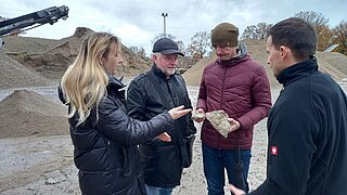 Vier Personen stehen im Kreis und blicken auf Steine, die eine Person in der Hand hält. im Hintergrund Berge einer Recyclinganlage. 