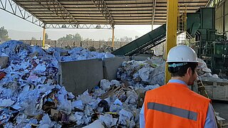 Ein überdachtes Recyclingzentrum mit großen Mengen sortierten Abfalls, darunter Papier und Plastik. Im Vordergrund steht eine Person mit weißer Schutzhelmkappe und orangefarbener Warnweste.