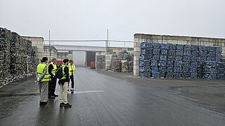 Eine Gruppe von vier Personen in leuchtend gelben Warnwesten steht auf dem Gelände einer Recyclinganlage. Im Hintergrund sind große Stapel gepresster Plastikballen zu sehen.