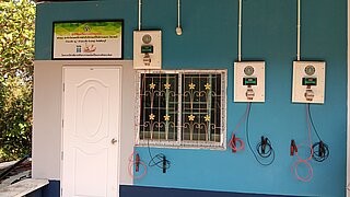 Eine blaue Hauswand, an der drei elektrische Ladesäulen mit jeweils zwei Ladekabeln befestigt sind. Über der Tür, die sich links an der Hauswand befindet, hängt ein Schild mit thailändischer Schrift.