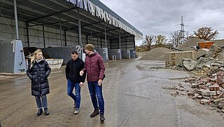 Drei Personen laufen über Fabrikgelände. Im Hintergrund befinden sich Bauschutt und Container. 