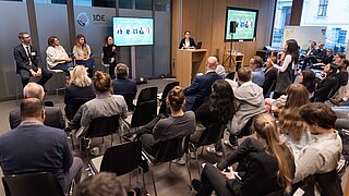 Ein Konferenzraum mit einer Podiumsdiskussion. Vier Personen sitzen links auf Barhockern und diskutieren, während eine Frau am Rednerpult rechts spricht.