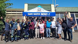 Eine Gruppe von Menschen steht jubelnd und sich freuend vor einem Pavillon mit der Aufschrift "Wasser sparen jetzt!"