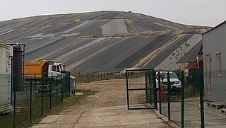 Eine grosse, abgedeckte Deponie mit dunklen Schutzplanen, die in Streifen angeordnet sind. Im Vordergrund stehen ein orangefarbener Lastwagen, ein Zaun, ein Tor und mehrere Container.