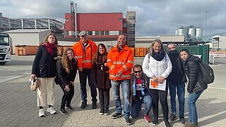 Gruppenfoto von neun Personen, die auf einem Fabrikgelände stehen. 