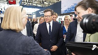 Messehalle mit mehreren Personen in geschäftlicher Kleidung. Im Vordergrund reicht eine Person mit blonden Haaren einer anderen Person die Hand. Im Hintergrund sind Messestände, eine große weiße Wand mit blauem Motiv und ein rotes Schild mit der Aufschrift 'B2' zu sehen. Die Szene wirkt geschäftig und professionell.