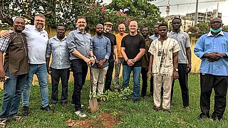 Eine Gruppe von 13 Männern steht um einen frisch eingepflanzten Baum