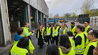 Eine Gruppe von Personen in gelben Warnwesten steht vor einer Fabrikhalle und hört einem Mann zu, der etwas erklärt. 