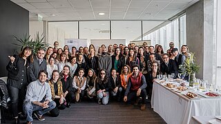 Gruppenfoto einer größeren Personengruppe, die in einem Veranstaltungsraum aufgereiht neben- und hintereinadersteht. Die erste Reihe hockt. Am rechten Rand steht ein Buffet.