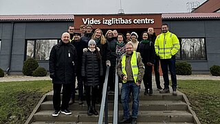 Eine Gruppe von 16 Personen steht auf einer Treppe vor einem Gebäude mit einem großen Schriftzug über dem Eingang. 