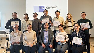 Ein Gruppenfoto von 12 Personen, wovon die Hälfte auf Stühlen sitzt und die andere Hälfte dahinter steht. Mehrere Personen halten ein Zertifikat in der Hand. Im Hintergrund ist eine Folie mit der Aufschrift "IHK Düsseldorf" zu sehen. 