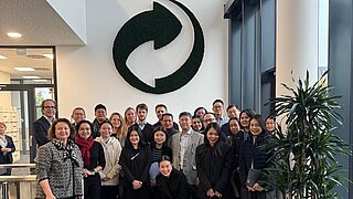 Gruppenfoto von mehreren Personen in einem Foyer. Über der Gruppe ist ein großes rundes Logo an der Wand zu sehen mit zwei ineinander verwobenen Pfeilen. 